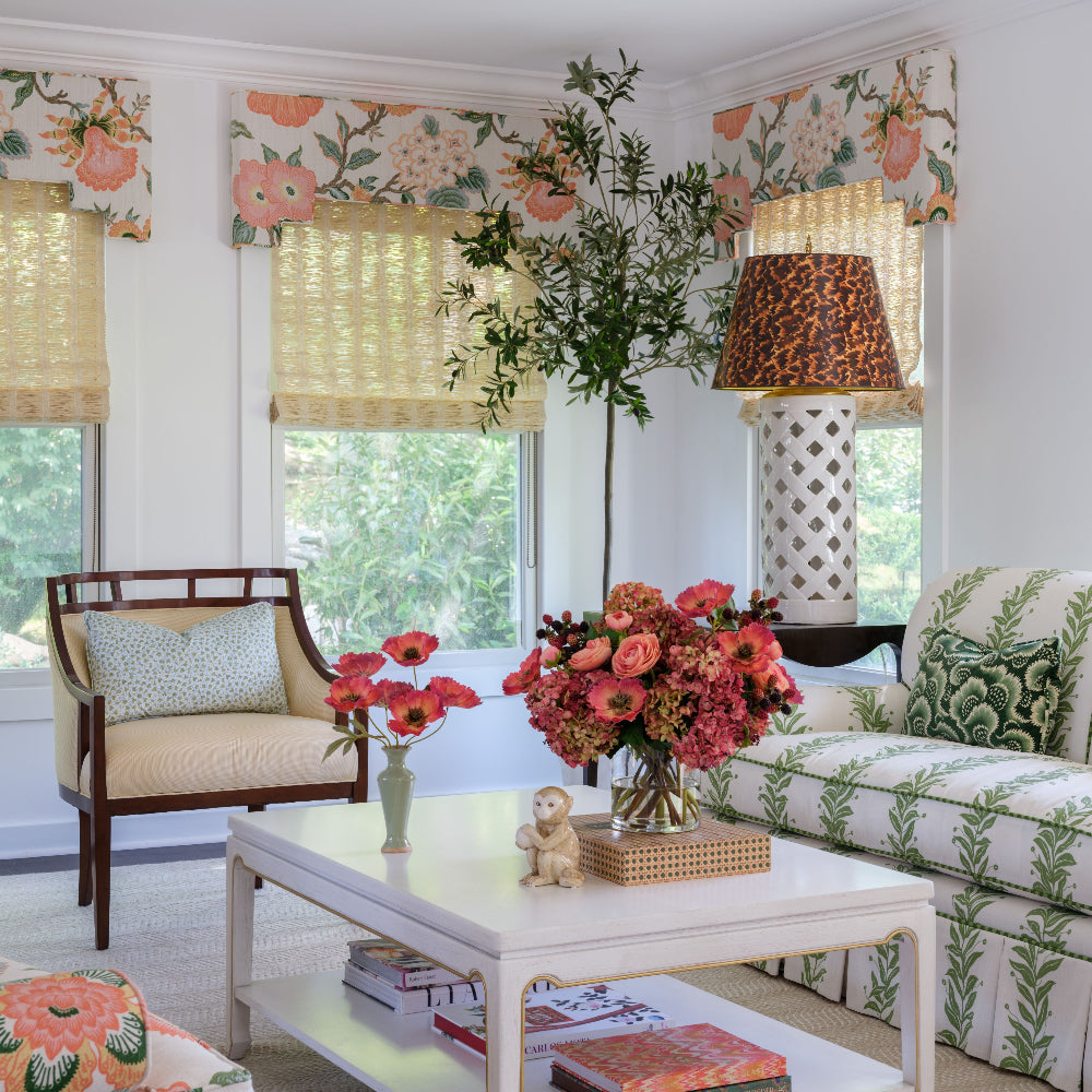A bright, styled living room featuring a white sofa with a green botanical pattern, a matching armchair, and floral window valances and pillows. A white coffee table with Diane James Home faux floral bouquets sits in the foreground, and a patterned lamp stands near a tall plant.