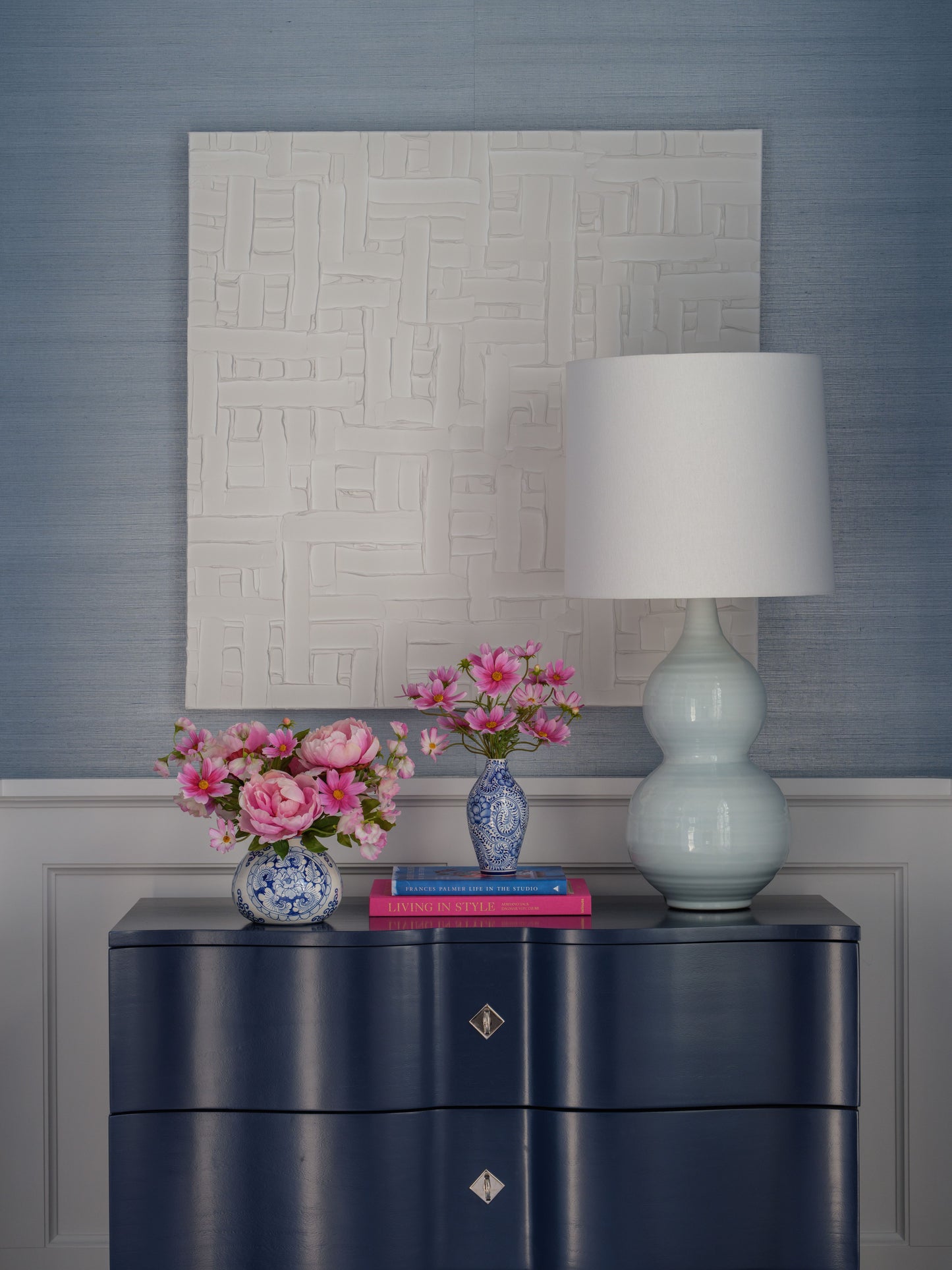 Navy blue dresser with a lamp, books, and faux floral arrangements by Diane James Home against a textured wall.