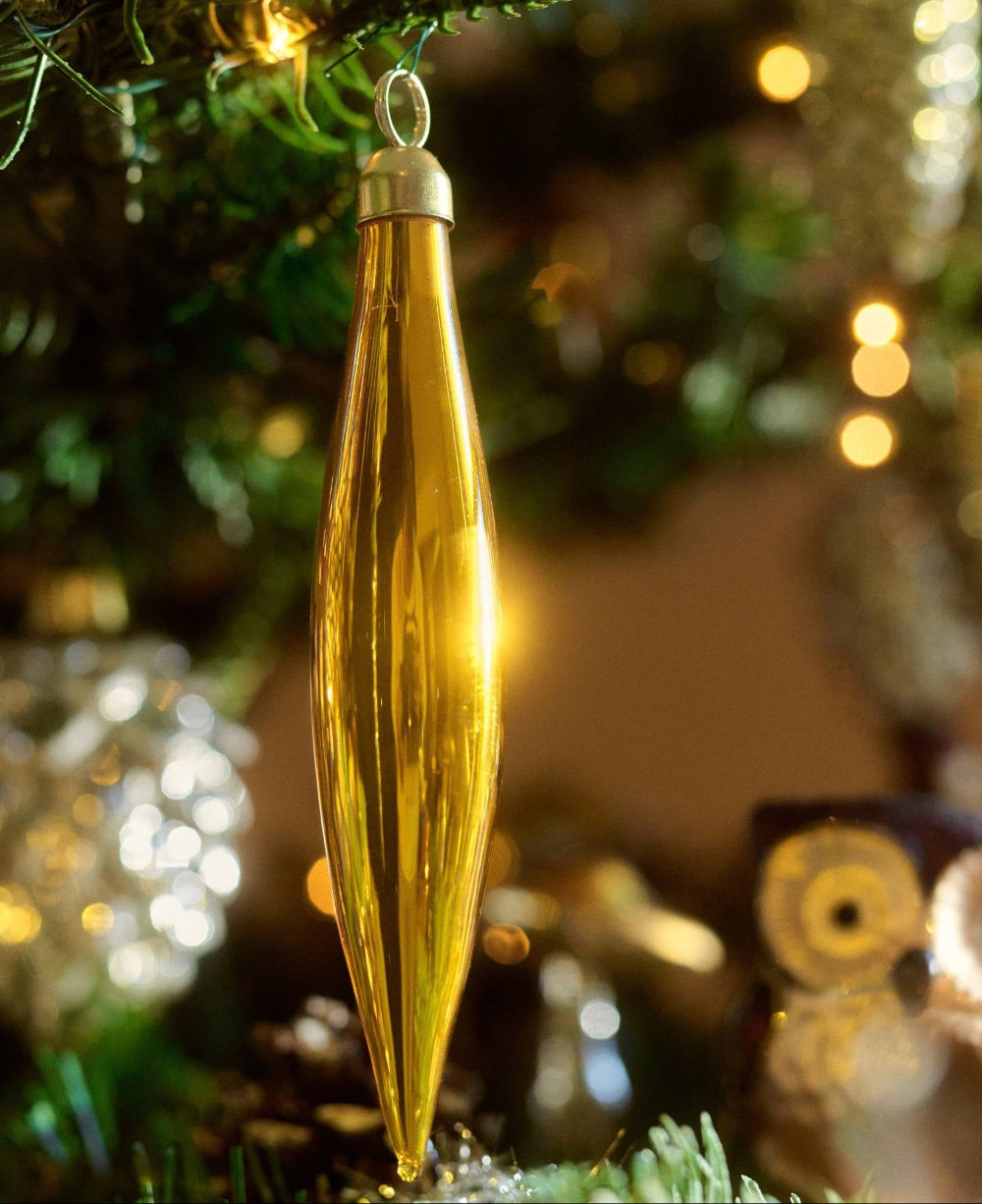 A shiny gold Christmas ornament shaped like a teardrop or leaf hangs from a decorated Christmas tree branch, with warm bokeh lights in the background.