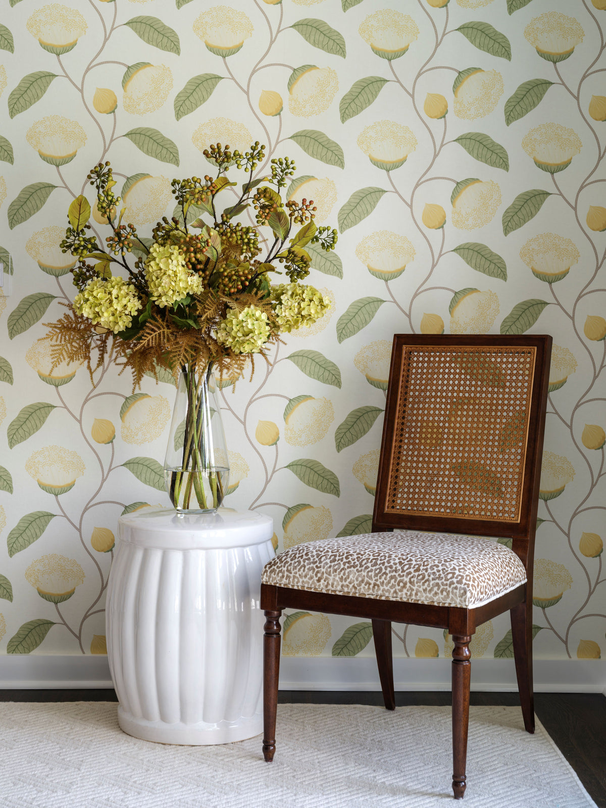 A styled interior corner featuring a wooden dining chair with a beige and brown animal-print cushion, set next to a white ribbed pedestal table holding a large pale green and brown faux floral arrangement by Diane James Home. The wall is covered in a light cream wallpaper with a repeating pattern of yellow lemons and green leaves.