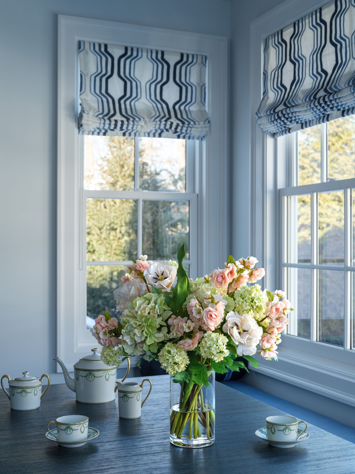 Floral arrangement on a table with teacups and teapots in a room with striped window treatments.