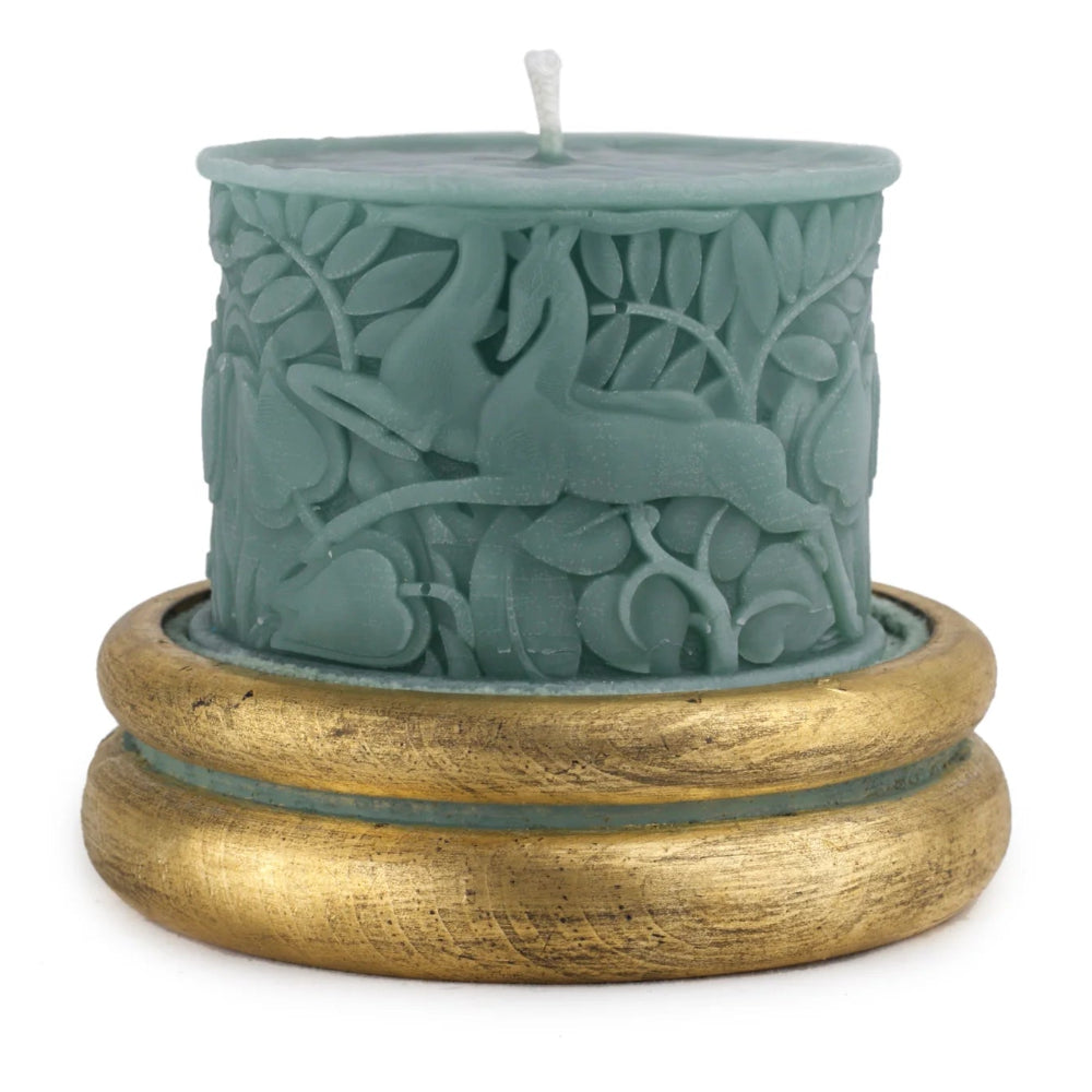 A sage green pillar candle with a detailed deer and foliage relief design, resting on a wide, textured gold-colored base against a white background.