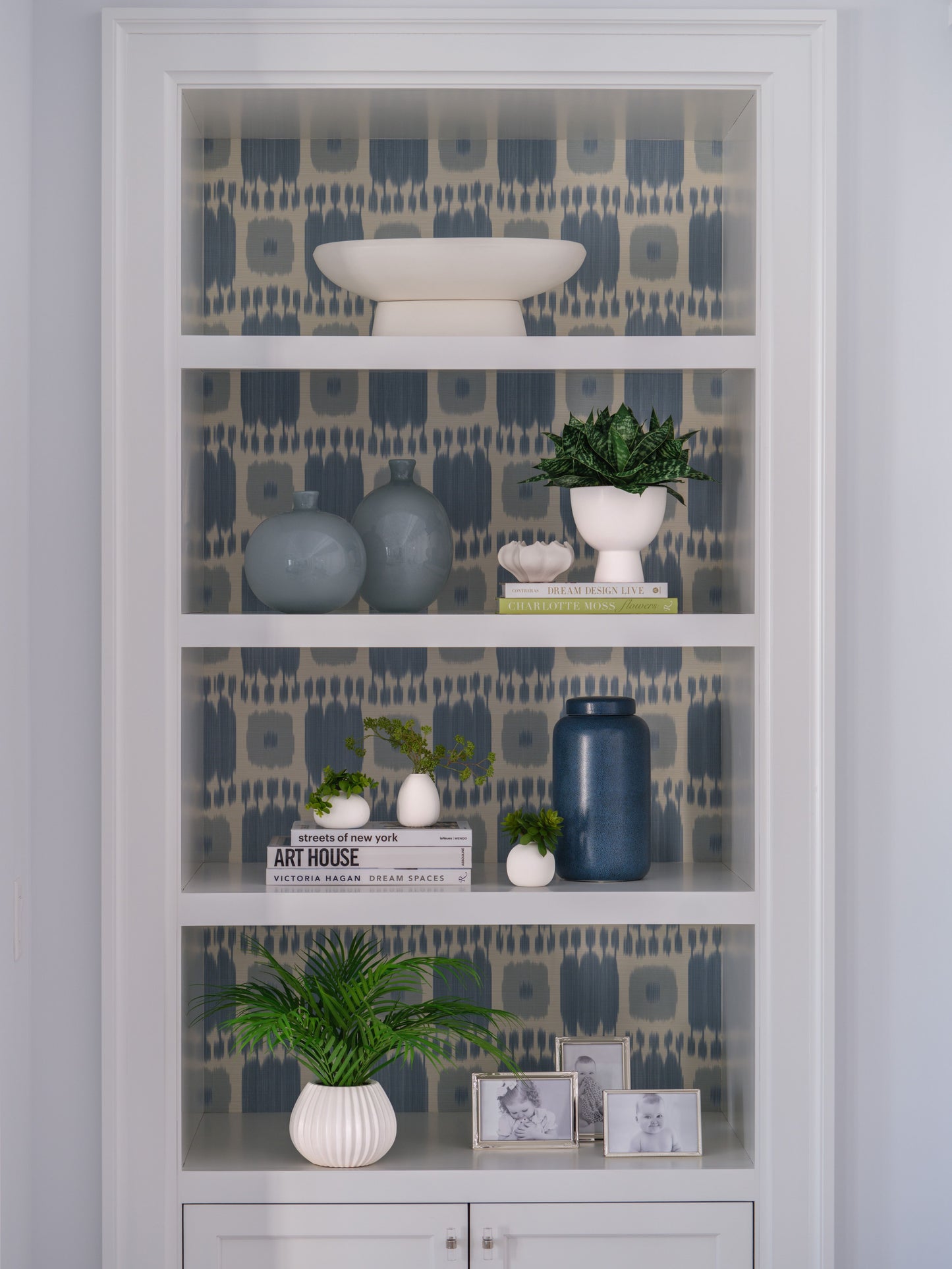 Decorative bookshelf with patterned wallpaper behind, featuring vases, books, and faux plants by Diane James Home.