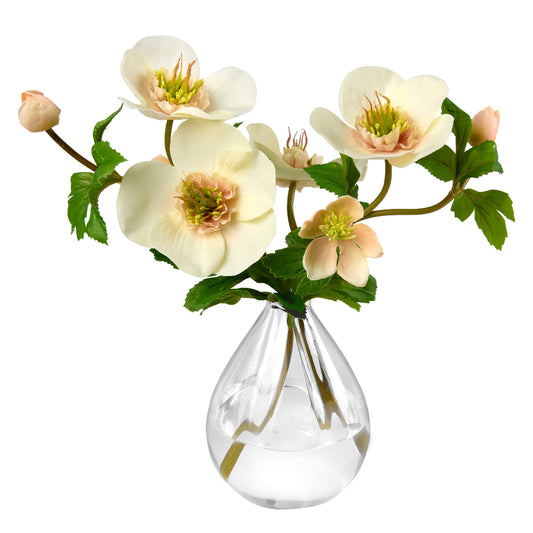 A bouquet by Diane James Home of artificial hellebores in a clear glass vase, featuring cream and peach blossoms with green leaves, set against a plain white background.
