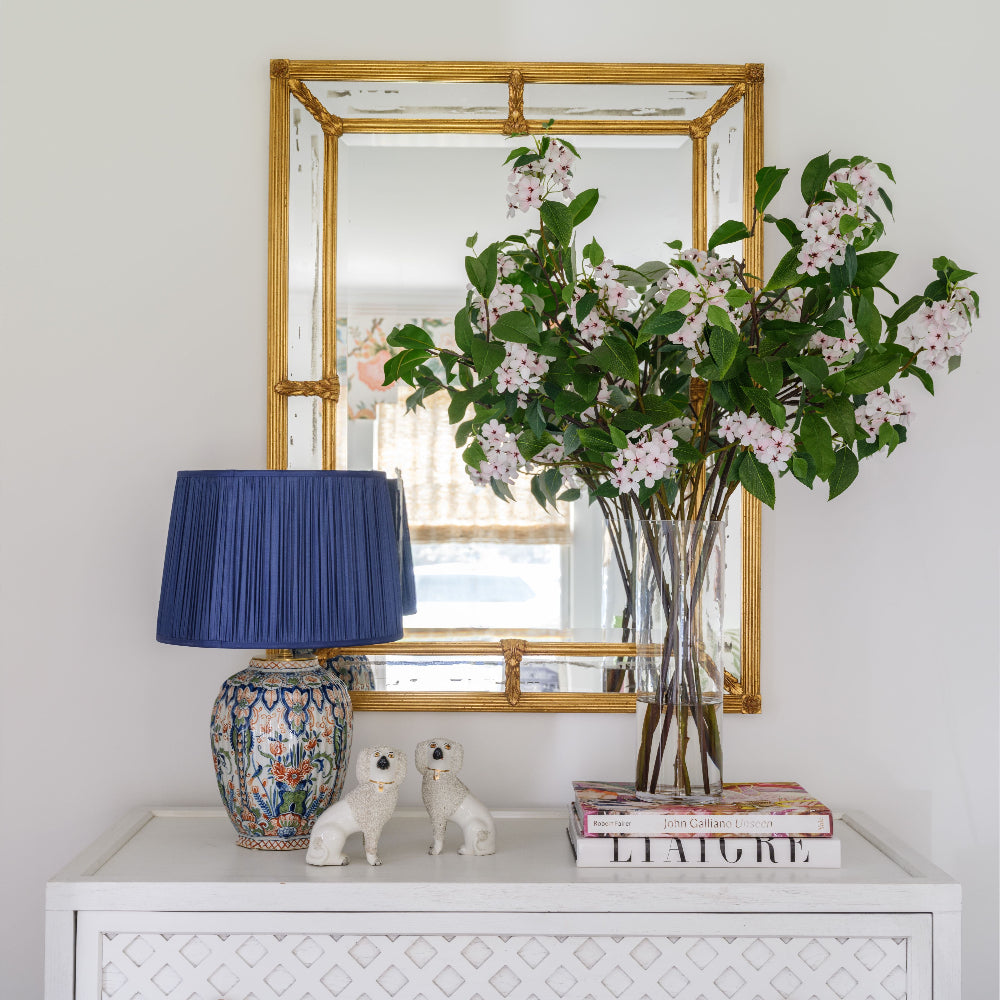 A white dresser with a geometric pattern and brass knobs, styled with a blue pleated lamp, ceramic dog figurines, books, and a large glass vase filled with a luxury artifical green leaves and pale pink blossoms by Diane James Home. A gold-framed mirror hangs on the white wall above.