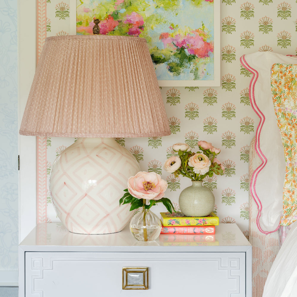 A styled bedroom corner featuring a white nightstand with a pleated blush pink lamp, a small vase with faux arrangements  by Diane James Home, books, and a framed floral painting on a wall with patterned wallpaper.