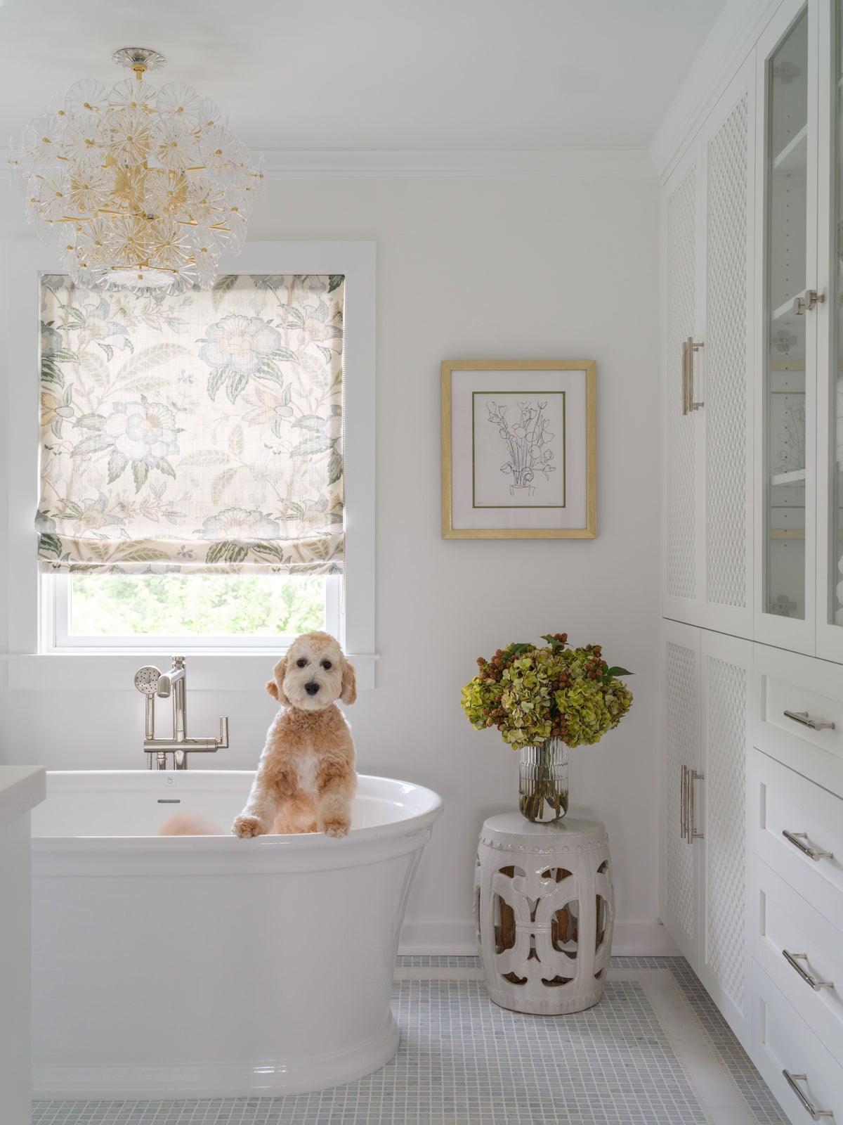 A fluffy light brown dog sits in a white freestanding bathtub in a bright, white bathroom. A floral-patterned roller blind is drawn over a window on the left, and a white stool with cutout details holding a vase of faux green hydrangeas by Diane James Home stands on the floor to the right.