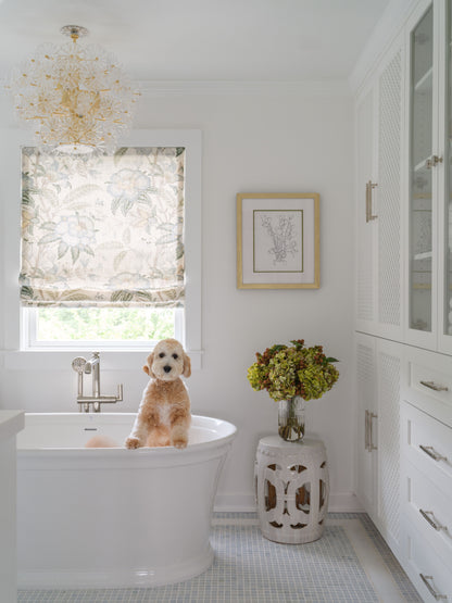 A fluffy light brown dog sits in a white freestanding bathtub in a bright, white bathroom. A floral-patterned roller blind is drawn over a window on the left, and a white stool with cutout details holding a vase of faux green hydrangeas by Diane James Home stands on the floor to the right.