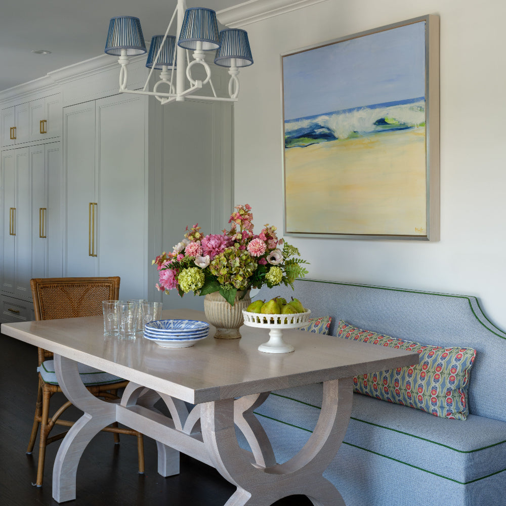 A styled dining room corner featuring a light wood table with sculptural white legs, a blue upholstered bench with patterned pillows, a luxury faux floral centerpiece by Diane James Home called Caledonia, and a framed beach painting on the wall.