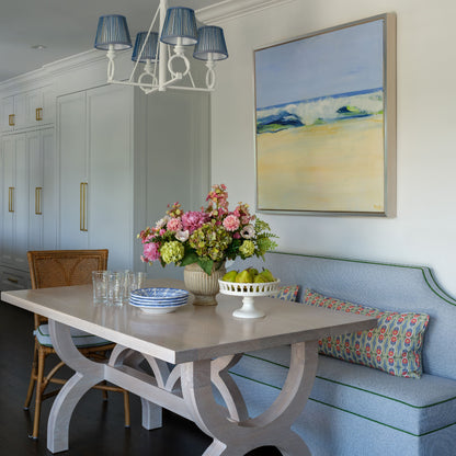 A styled dining room corner featuring a light wood table with sculptural white legs, a blue upholstered bench with patterned pillows, a luxury faux floral centerpiece by Diane James Home called Caledonia, and a framed beach painting on the wall.