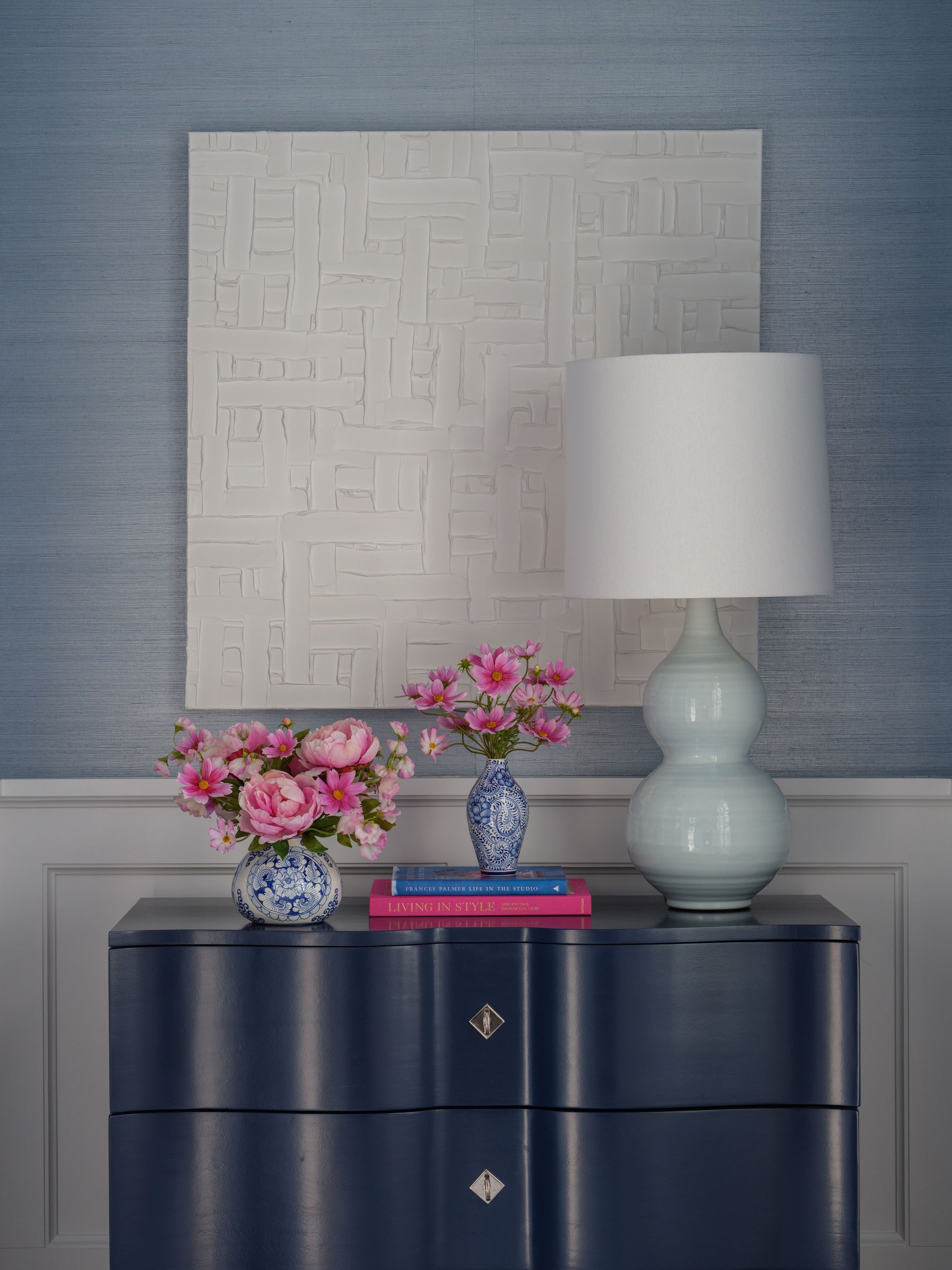 Navy blue dresser with a lamp, books, and faux floral arrangements by Diane James Home against a textured wall.