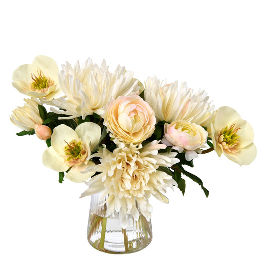 An artificial flower bouquet in a clear ribbed glass vase by Diane James Home featuring cream chrysanthemums, blush ranunculus, and white hellebores with green leaves, set against a plain white background.