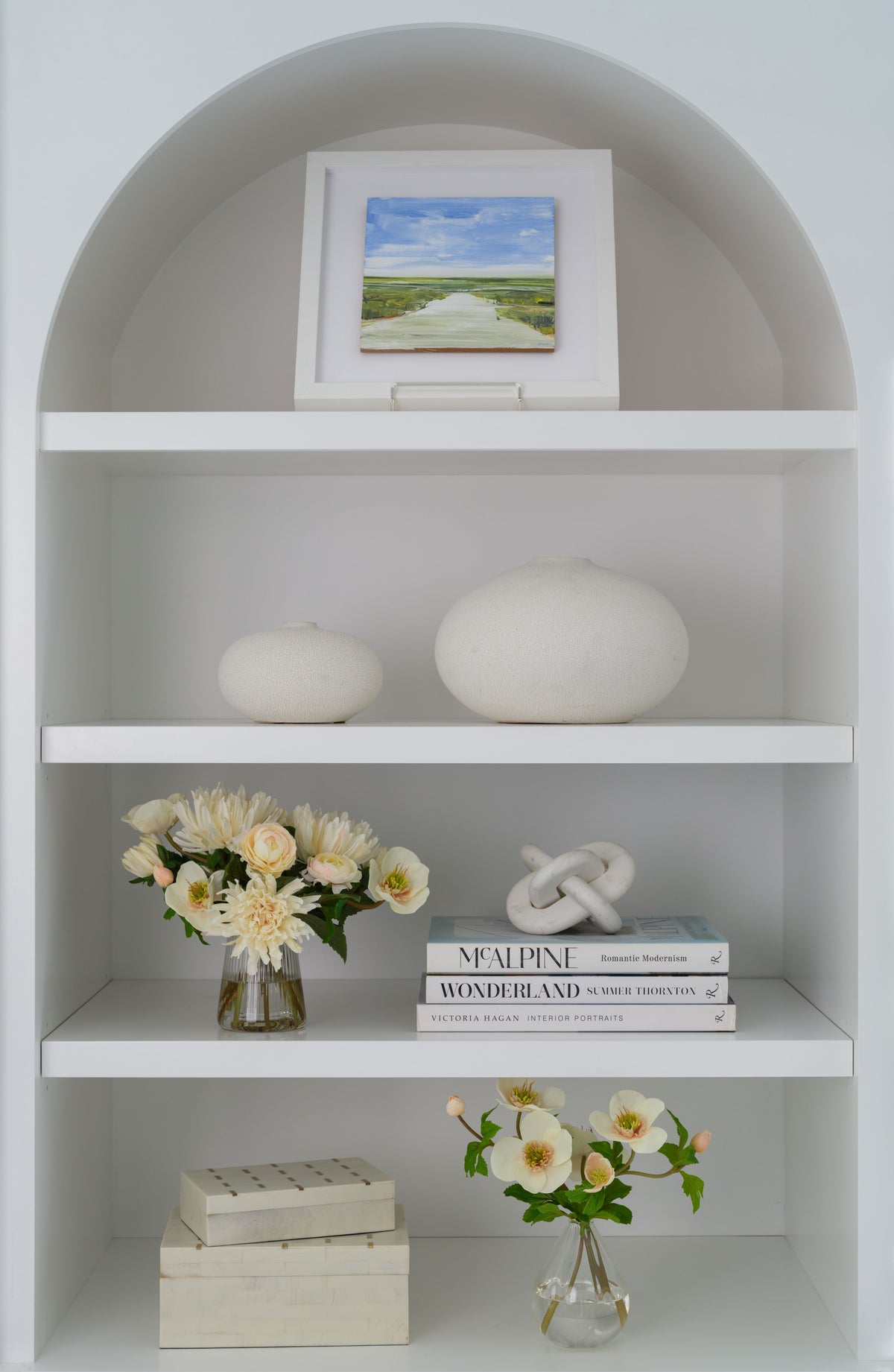 A white arched built-in shelf display with decorative objects including a small framed landscape print, two white ceramic vessels, a vase of faux cream flowers by Diane James Home, stacked books, and another  faux flower arrangement by Diane James Home.