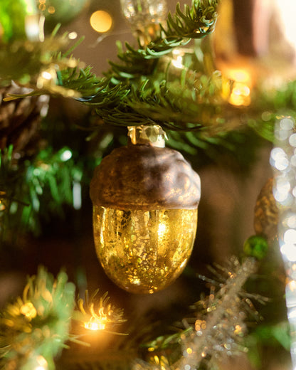 A close-up of a gold acorn-shaped Christmas ornament by Eddie Ross hanging on a green Christmas tree branch with warm bokeh lights in the background.