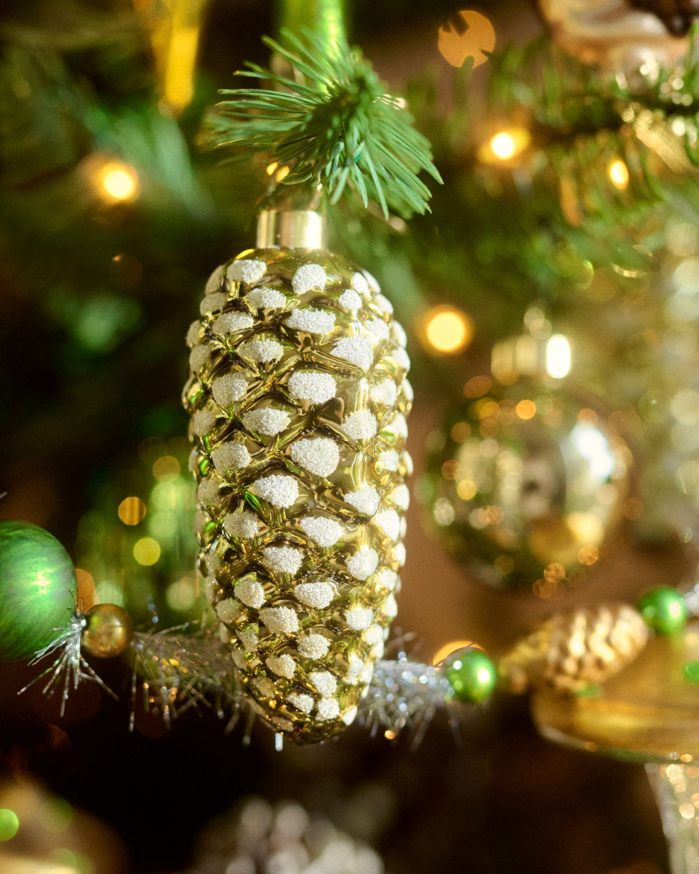 A close-up of a gold pinecone-shaped Christmas ornament covered in white glitter, hanging on a decorated Christmas tree with warm lights and green ornaments in the background.