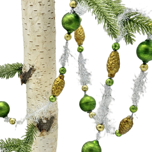 Green and gold Christmas tree garland with shiny green beads, textured gold pinecones, and silver tinsel draped over pine branches and a birch tree trunk on a white background.