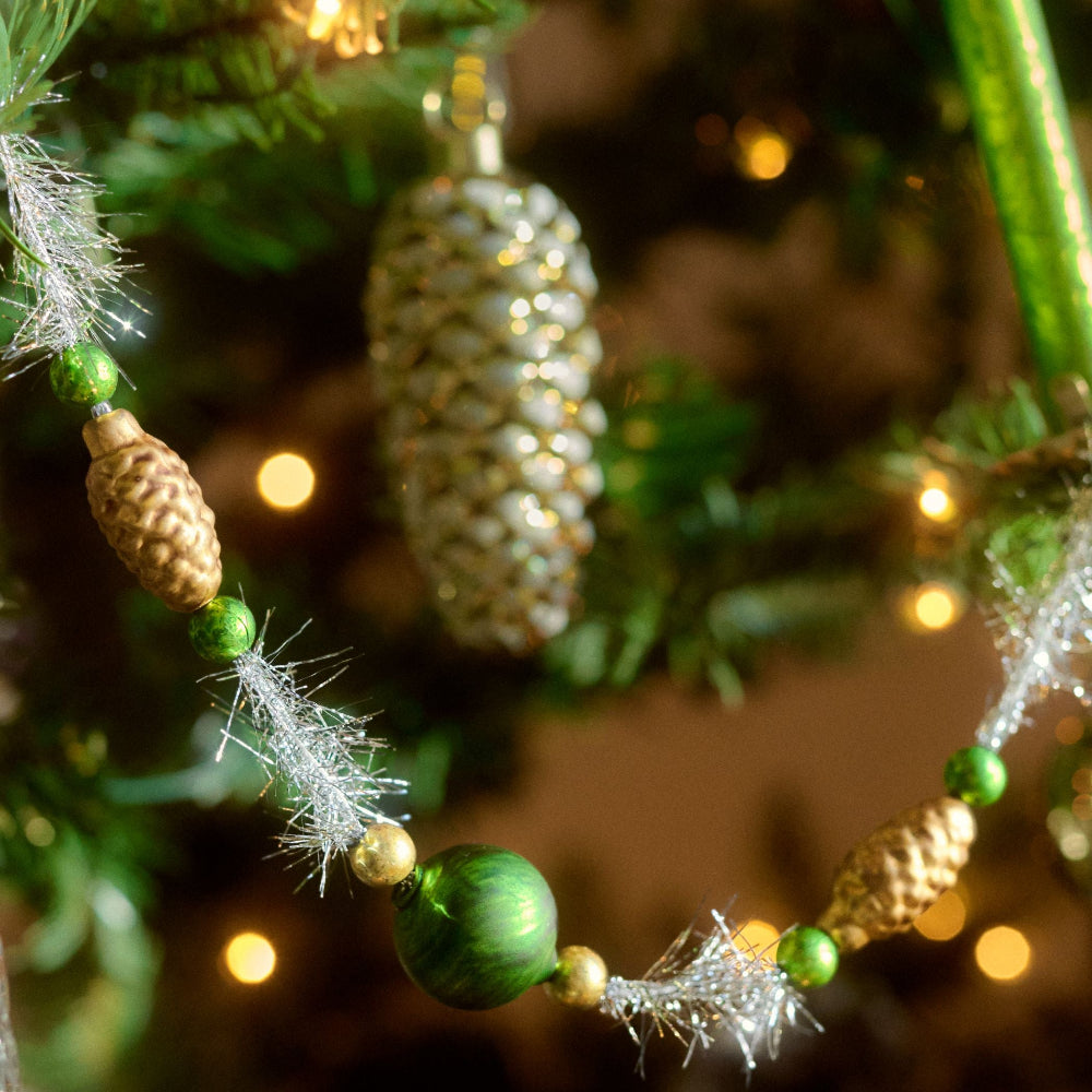 A close-up of a Christmas tree decoration garland featuring green and gold beads, miniature pinecones, and silver tinsel, draped over green pine branches with warm bokeh lights in the background.