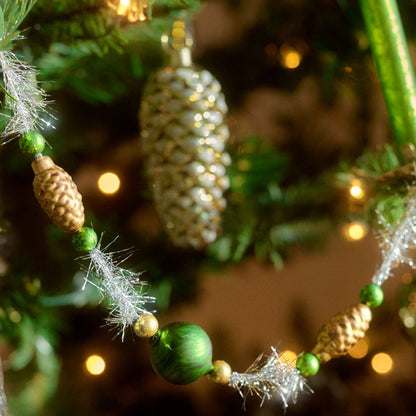 A close-up of a Christmas tree decoration garland featuring green and gold beads, miniature pinecones, and silver tinsel, draped over green pine branches with warm bokeh lights in the background.