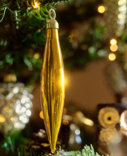 A shiny gold Christmas ornament shaped like a teardrop or leaf hangs from a decorated Christmas tree branch, with warm bokeh lights in the background.