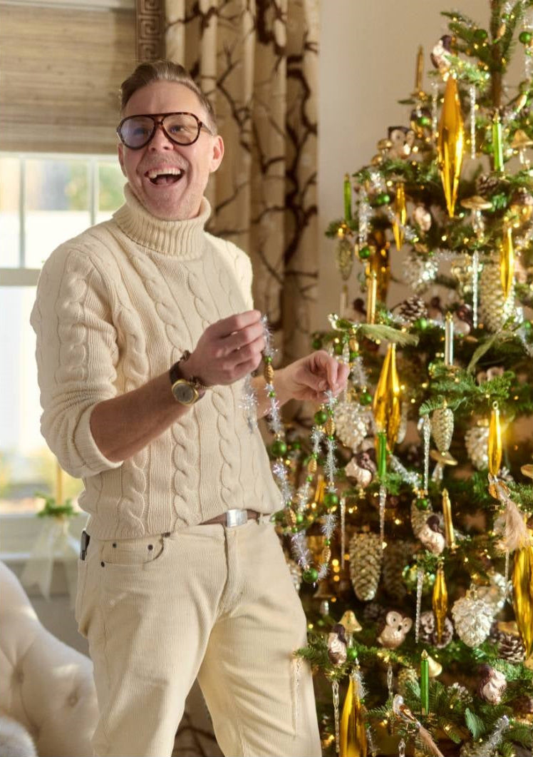 A man wearing a cream cable-knit turtleneck sweater and matching cream trousers stands next to a decorated Christmas tree, holding a string of ornaments in a warmly lit indoor setting.
