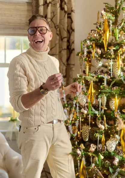 A man wearing a cream cable-knit turtleneck sweater and matching cream trousers stands next to a decorated Christmas tree, holding a string of ornaments in a warmly lit indoor setting.