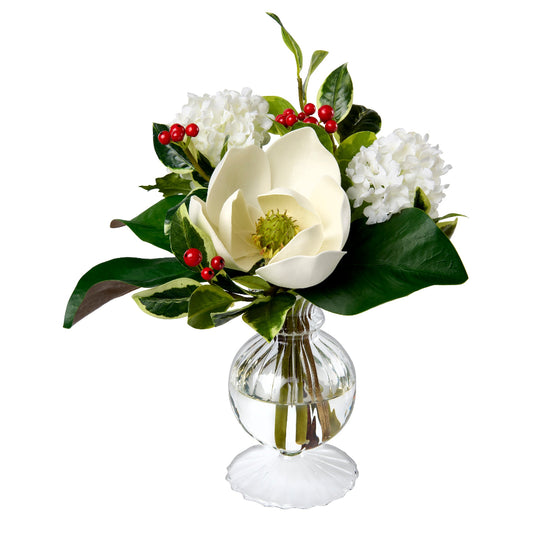 A faux floral arrangement by Diane James Home featuring a large white magnolia flower, white snowballs, green leaves, and holly with small red berries in a clear ribbed glass vase on a white background.