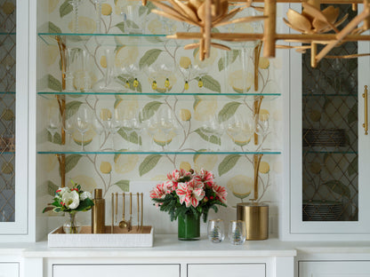 A styled bar area featuring a white cabinetry unit with open glass shelves displaying wine glasses, a floral wallpaper backdrop with yellow lemons and green leaves, a white countertop with a faux flower arrangement by Diane James, gold and glass accessories, and part of a wooden chandelier visible at the top.