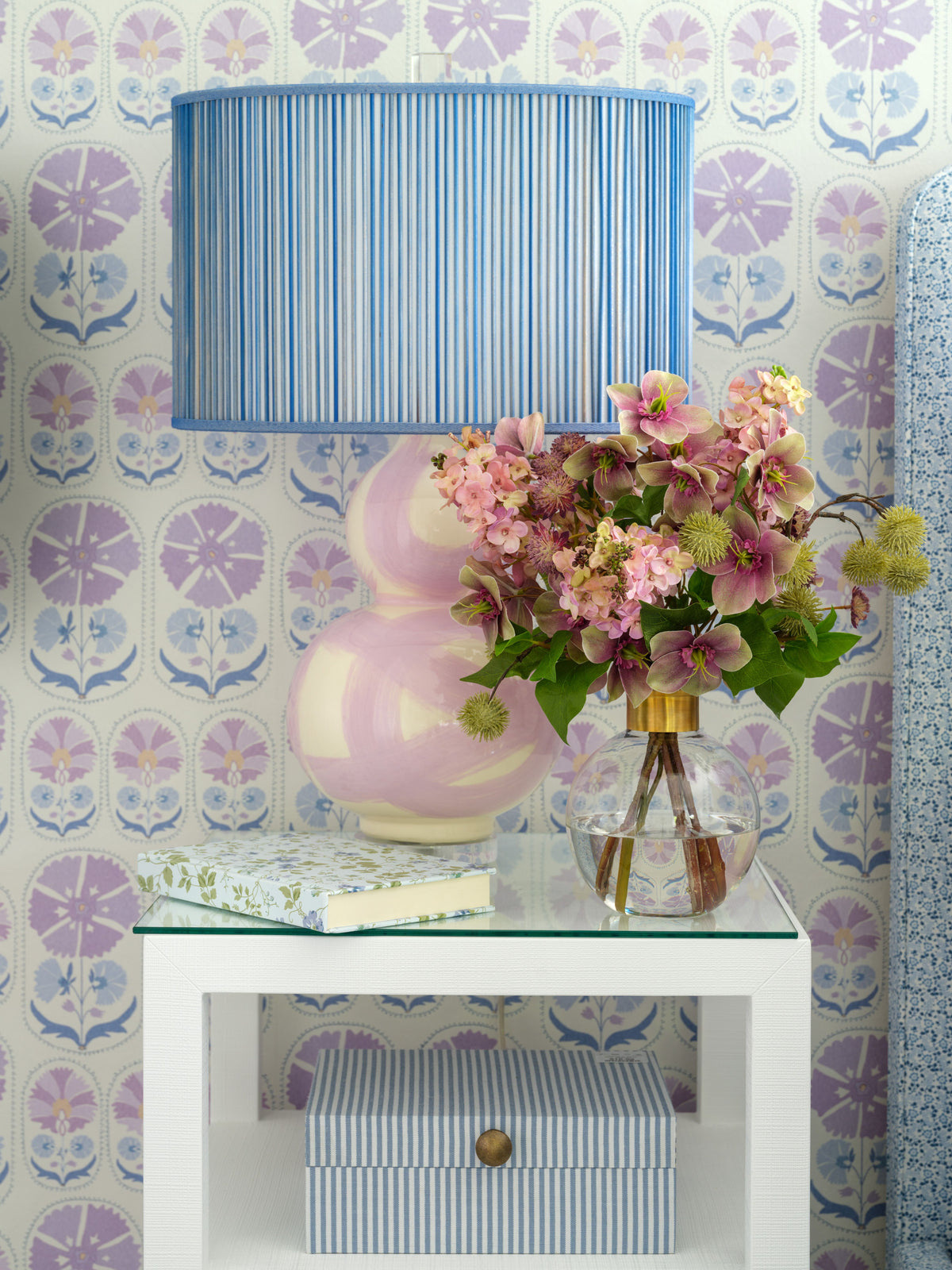 A styled interior scene showing a blue and white striped table lamp with a pink and white gourd-shaped base, placed on a white side table next to a glass vase of faux flowers by Diane James Home. The background features a purple and blue floral patterned wallpaper.