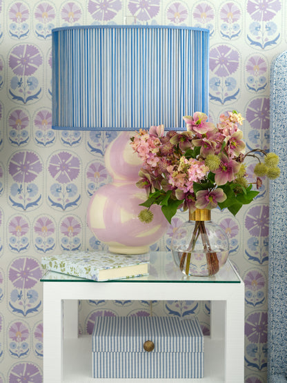 A styled interior scene showing a blue and white striped table lamp with a pink and white gourd-shaped base, placed on a white side table next to a glass vase of faux flowers by Diane James Home. The background features a purple and blue floral patterned wallpaper.