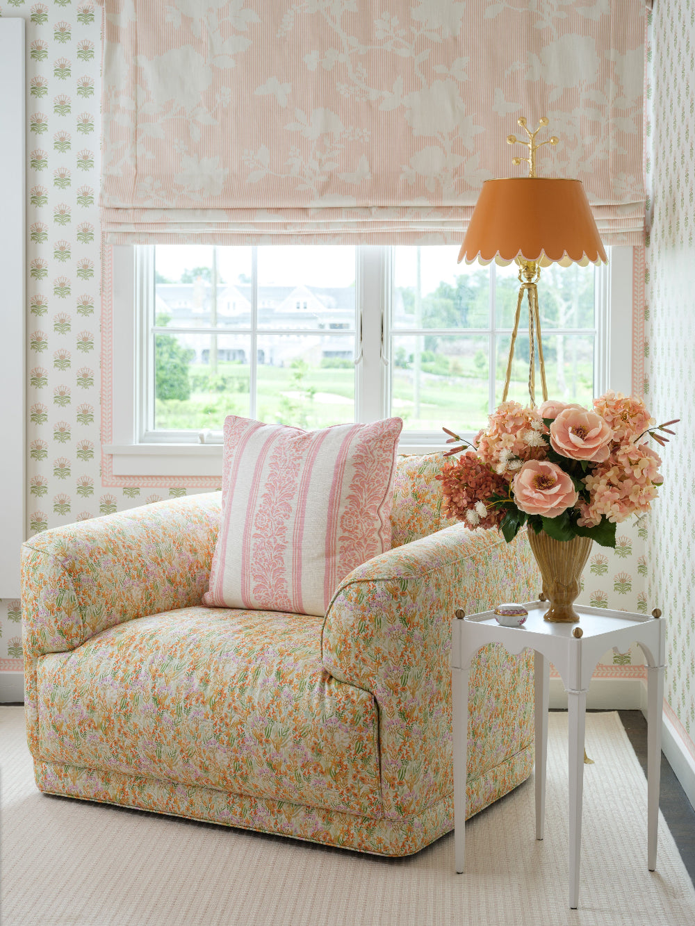 A cozy upholstered armchair in a light pink and yellow floral pattern sits beside a small white side table with Highland Fling, a luxury artificial bouquet by Diane James Home, and a gold lamp. A matching floral blind and patterned wallpaper frame a window in the background.