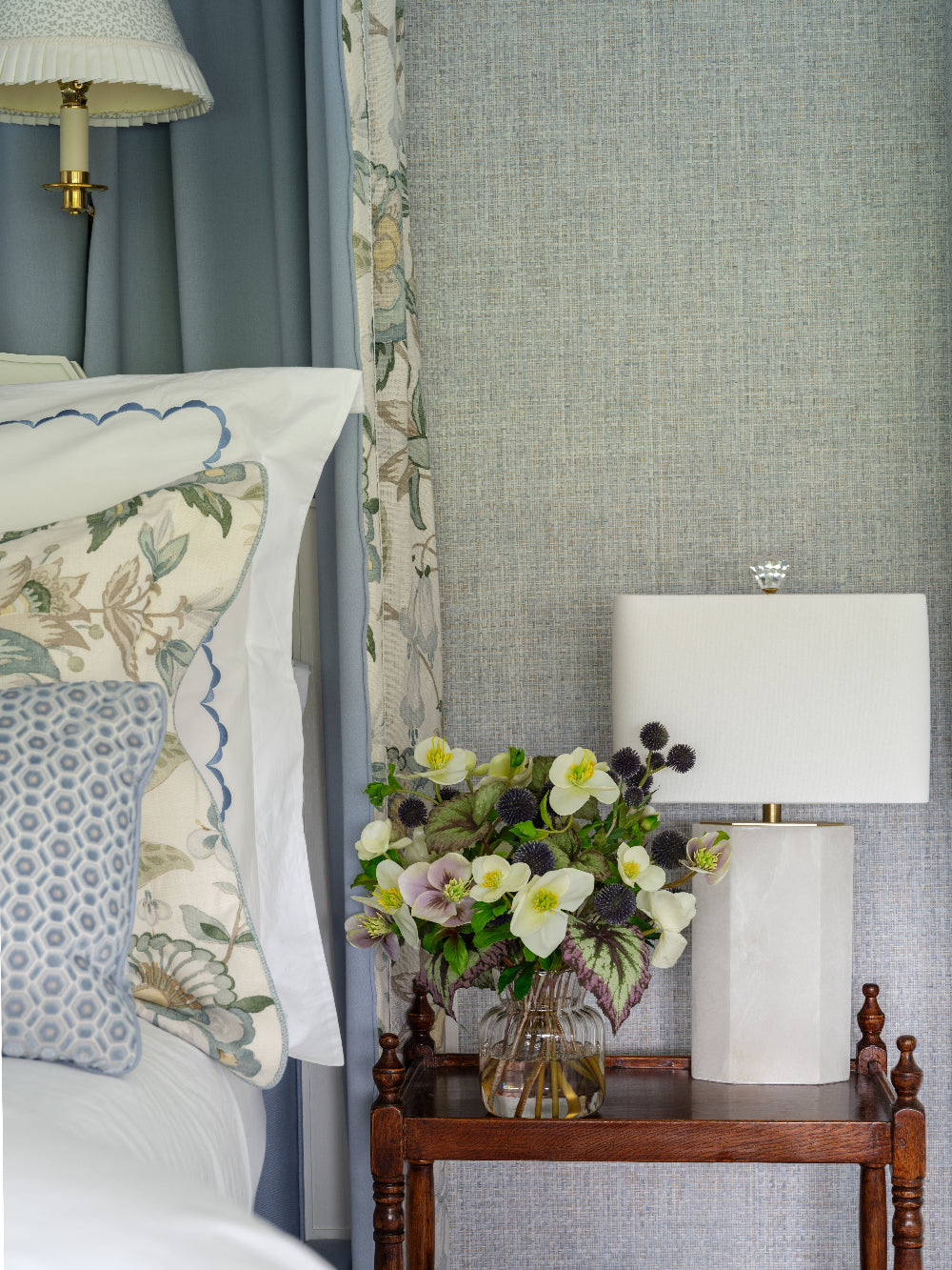 A styled bedroom corner featuring a wooden nightstand with a white table lamp, a glass vase of faux white and green flowers by Diane James Home, and a bed with white bedding, patterned pillows, and blue-gray curtains against a textured light blue-gray wall.