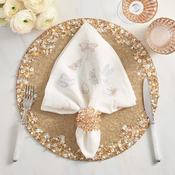 A styled place setting on a white marble surface featuring a round beige placemat with a gold and white beaded border, a white napkin with embroidered butterflies, a crystal-like napkin ring, silver flatware, and decorative glassware.