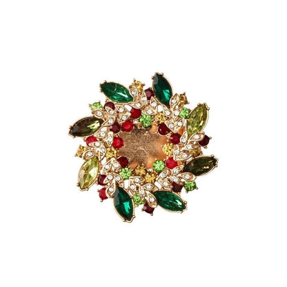 A round gold-tone napkin ring featuring a wreath of marquise-cut green, olive, and red gemstones, accented with small clear crystals on a white background.
