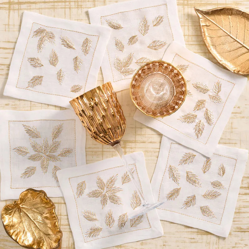 A styled flat-lay of white embroidered napkins with gold leaf motifs, arranged on a light woven surface with gold decorative leaf pieces and a gold textured glasses.