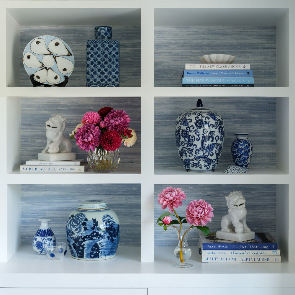 A white 3x3 shelving unit displaying various decorative blue and white porcelain vases, books, and floral arrangements by Diane James Home against a blue-gray wallpapered background.