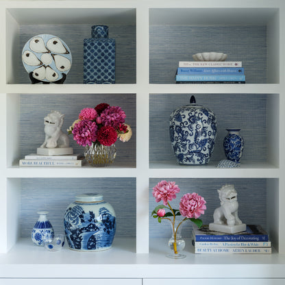A white 3x3 shelving unit displaying various decorative blue and white porcelain vases, books, and floral arrangements by Diane James Home against a blue-gray wallpapered background.