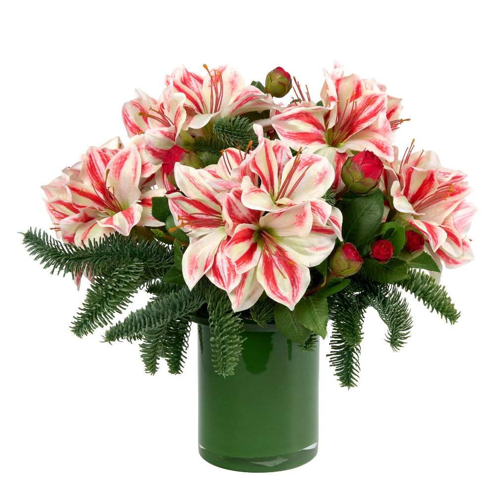 A faux floral arrangement by Diane James Home featuring large white amaryllis with red stripes, green pine, and small red camellia buds in a dark green cylindrical vase, photographed on a white background.