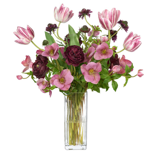 A vibrant artificial flower arrangement by Diane James Home featuring mauve hellebores, pink-and-white tulips, and dark burgundy ranunculus and scabiosa in a tall, clear glass vase, set against a solid white background.