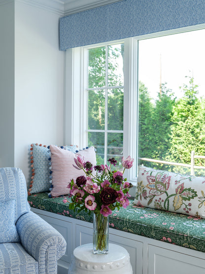 A bright window seat with a green floral cushion, patterned pillows, a blue patterned valance, and a vase of faux mauve and purple flowers by Diane James Home on a white side table.