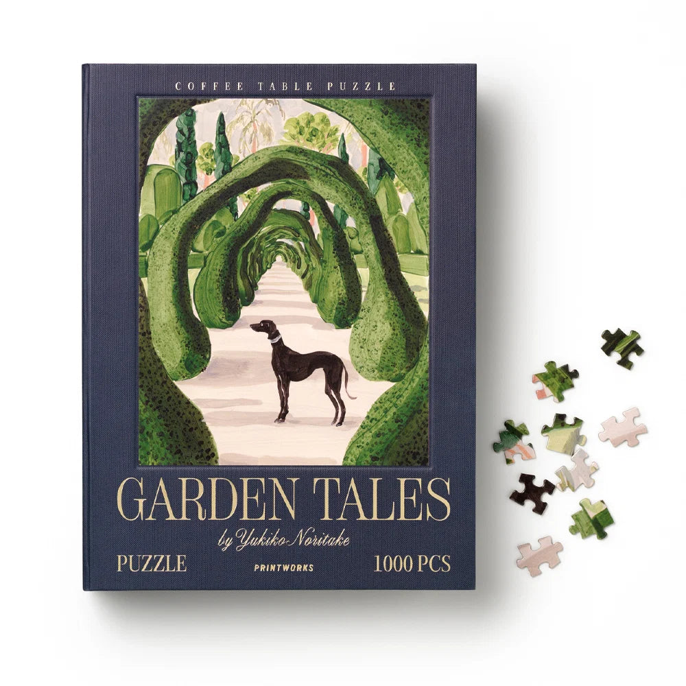 A 1000-piece coffee table puzzle titled 'Garden Tales' by Yoshiko Nishitake, shown as a dark navy blue book-style box with an illustrated garden archway scene. Several loose puzzle pieces are scattered to the right on a clean white background.