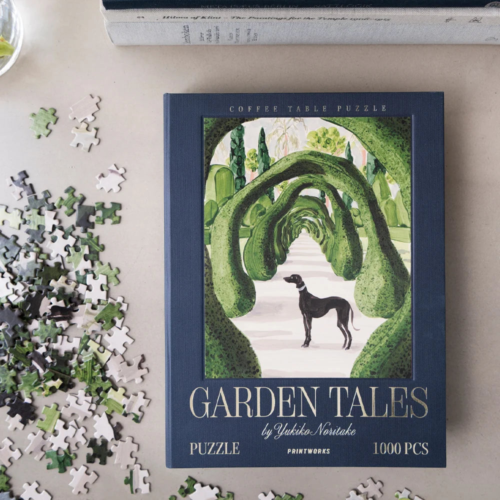 A completed and partially assembled coffee table puzzle titled 'Garden Tales' by Yoko Natorioka, displayed on a beige tabletop with loose puzzle pieces scattered around it.
