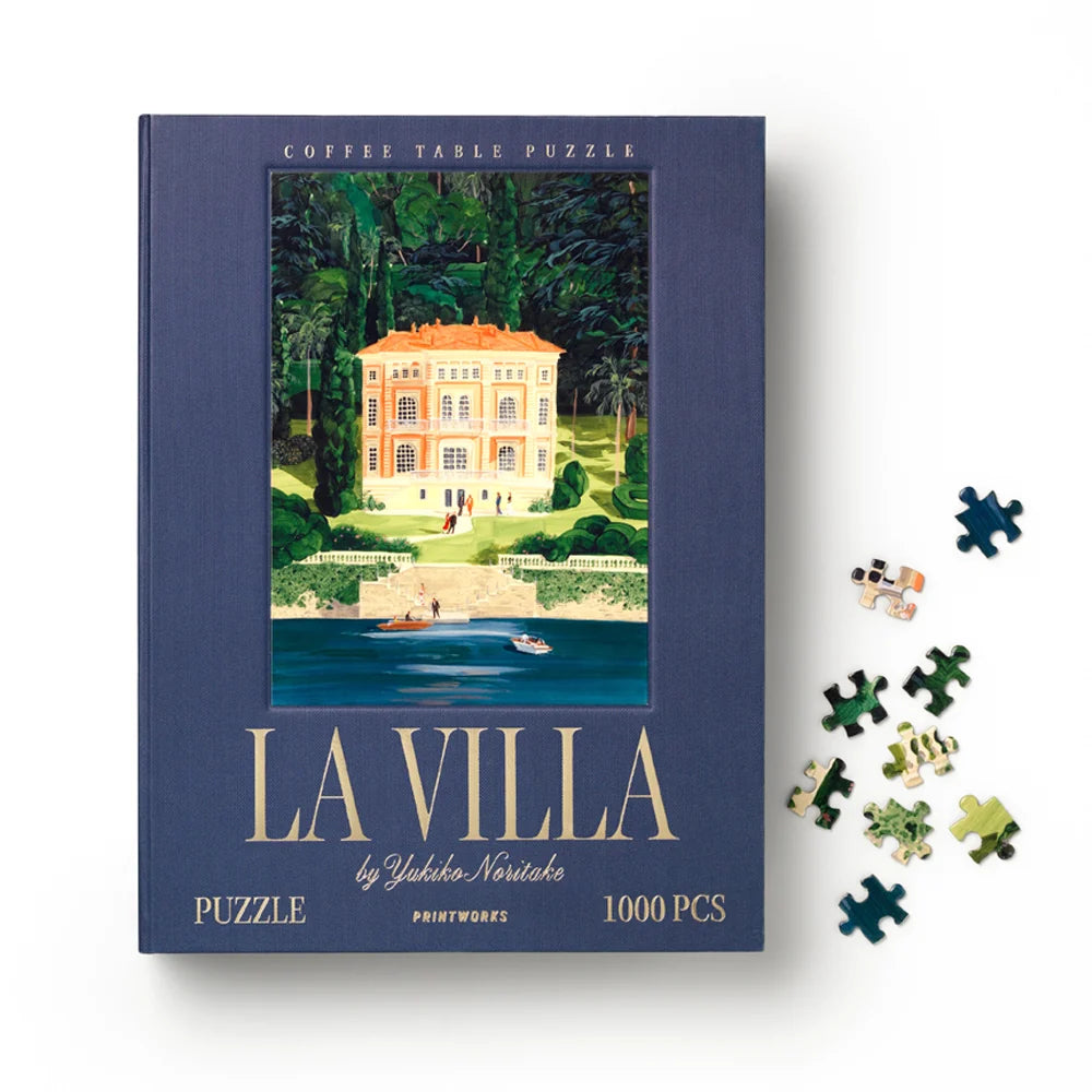 A coffee table puzzle book titled 'LA VILLA' by Yekaterina Nesterko, featuring a navy blue cover with an illustration of a villa by the water. Several loose puzzle pieces are scattered to the right on a white background.