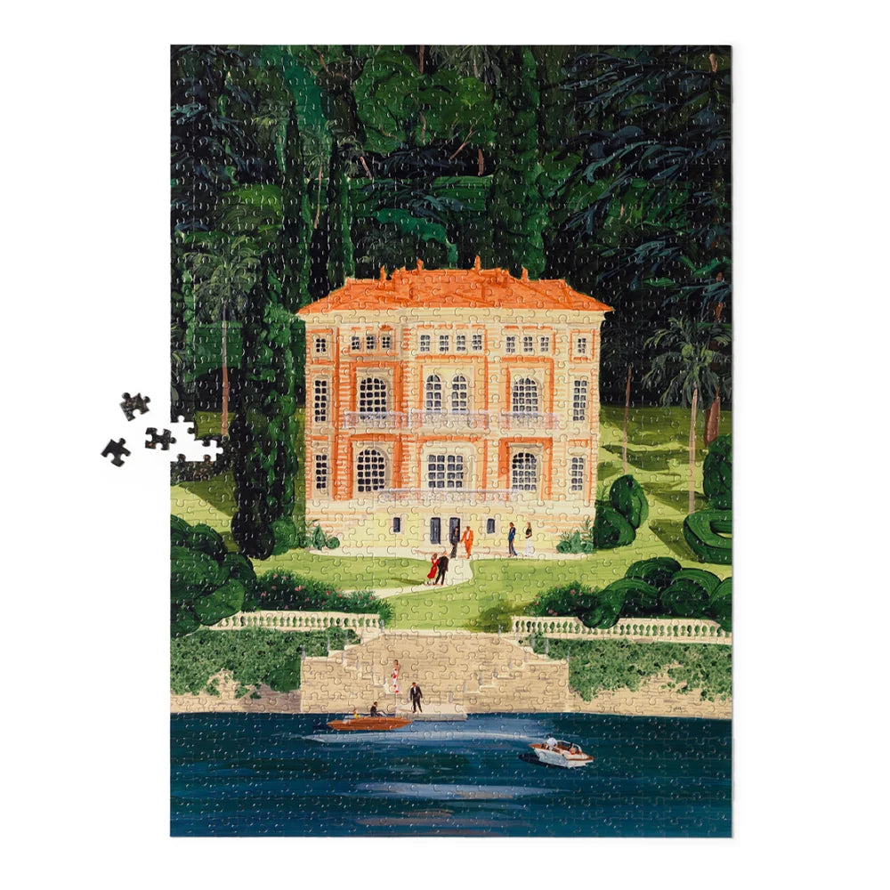 A completed jigsaw puzzle depicting an elegant orange-roofed villa on a green lawn beside a lake, with a few loose puzzle pieces floating to the left on a white background.
