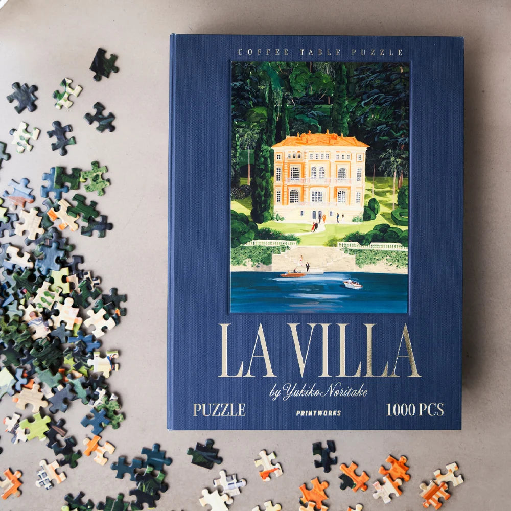 A coffee table puzzle box titled 'LA VILLA' by Yekaterina Nesterko, surrounded by scattered puzzle pieces on a neutral surface. The box is dark blue with an illustration of a villa by the water, and the puzzle contains 1000 pieces.