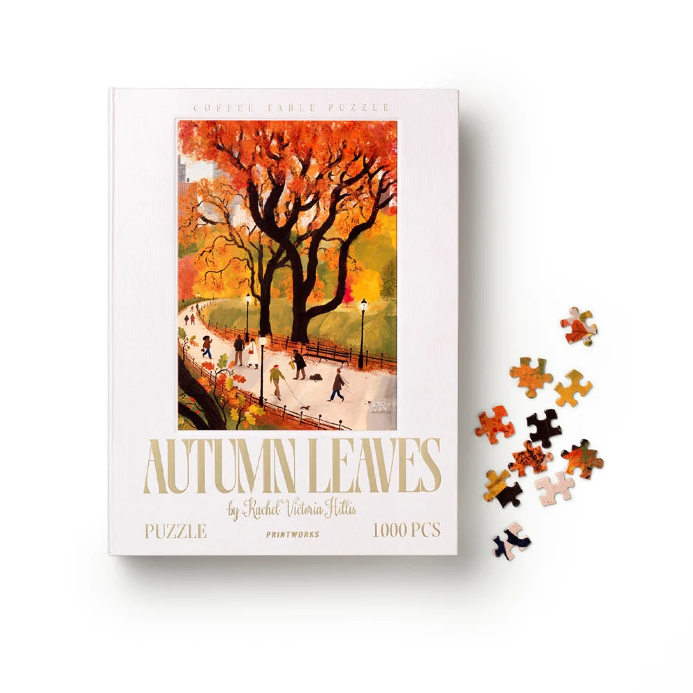 A jigsaw puzzle box titled 'Autumn Leaves' by Rachel Victoria Huttles, featuring a colorful illustration of a tree with orange and yellow leaves on a park path. Several loose puzzle pieces are scattered to the right on a clean white background.