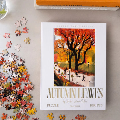 A 1000-piece coffee table puzzle titled 'Autumn Leaves' by Rachel Victoria Hells, displayed on a light gray surface with loose puzzle pieces scattered around it. The cover features an illustration of a large tree with vibrant orange and yellow autumn leaves, with people walking along a park path.