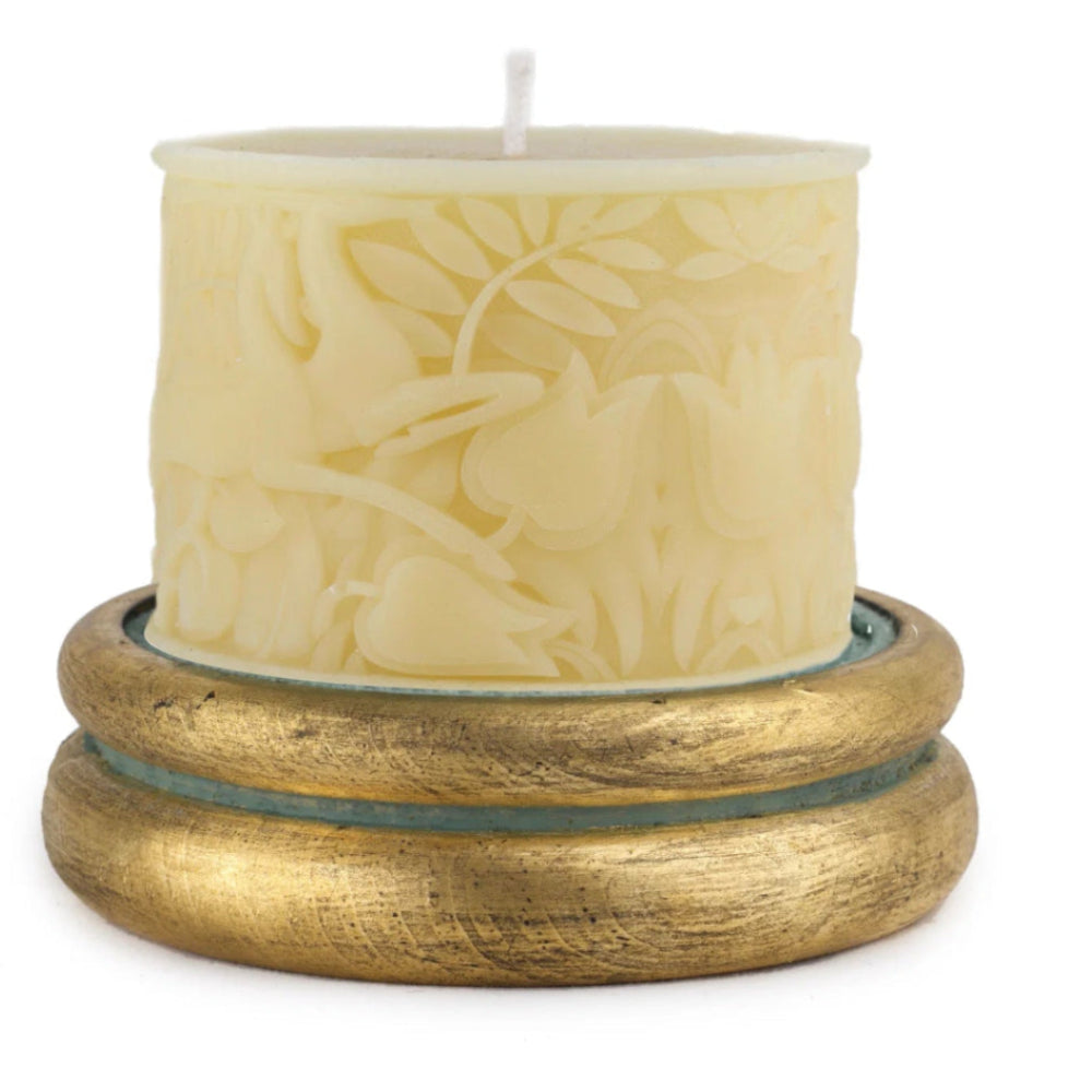 A cream-colored pillar candle with an embossed deer and floral pattern, resting on a textured gold-colored wooden base against a white background.