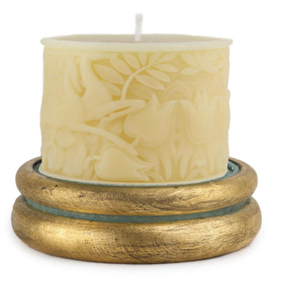 A cream-colored pillar candle with an embossed deer and floral pattern, resting on a textured gold-colored wooden base against a white background.