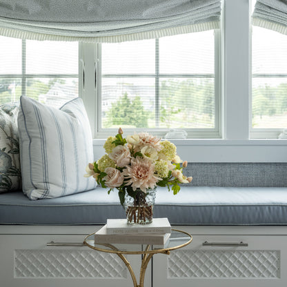 A styled interior window seat with light blue-gray Roman shades, matching cushions, striped pillows, and a small glass table holding a bouquet by Diane James Home of faux dhalias, hellebores, roses and viburnum in a hobnail vase.