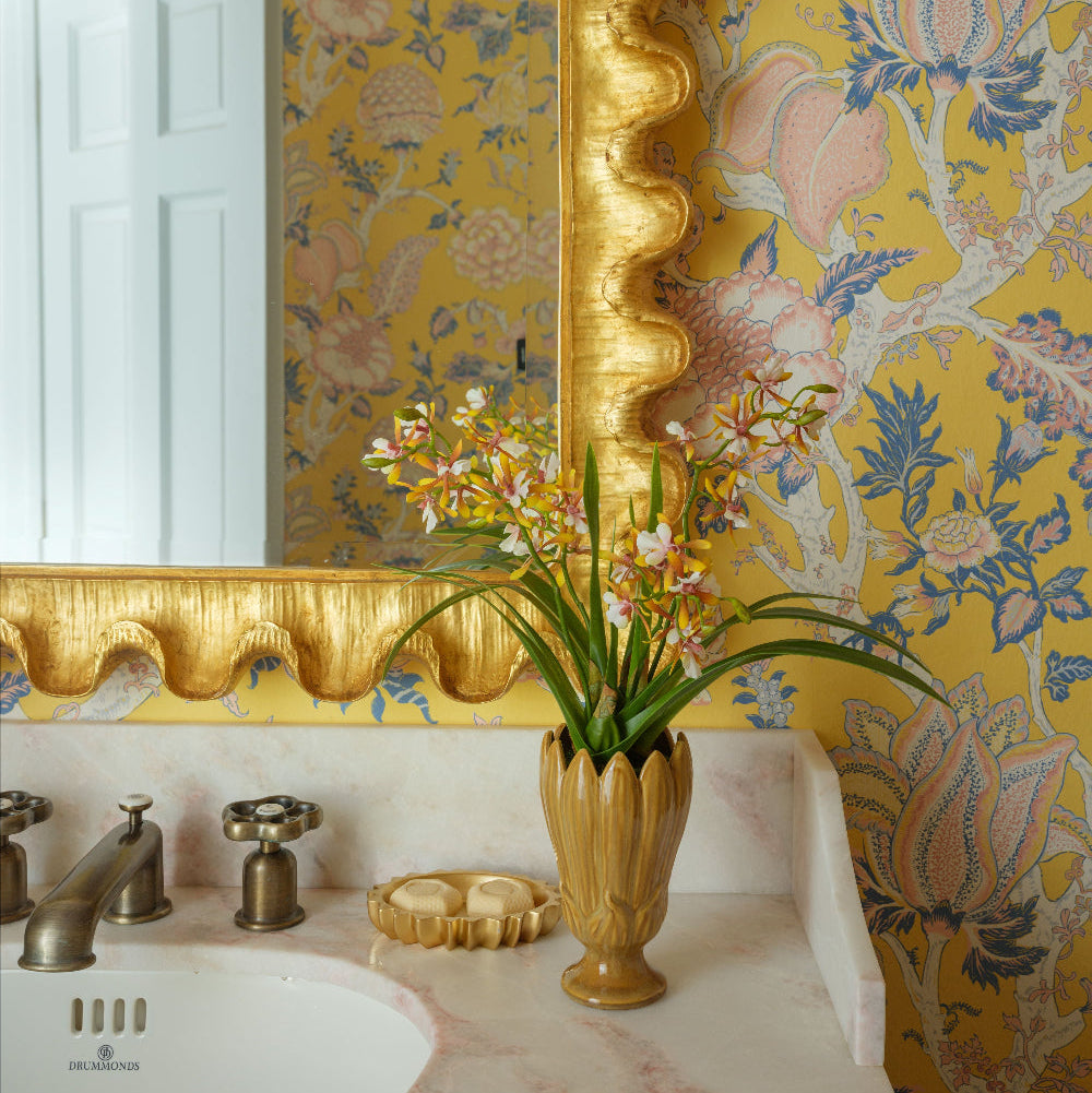 A styled bathroom corner featuring a white sink with a gold-toned faucet, a faux cattleya orchid in a ceramic vase by Diane James Home on a marble countertop, and a large mirror with a decorative gold frame against yellow floral wallpaper.
